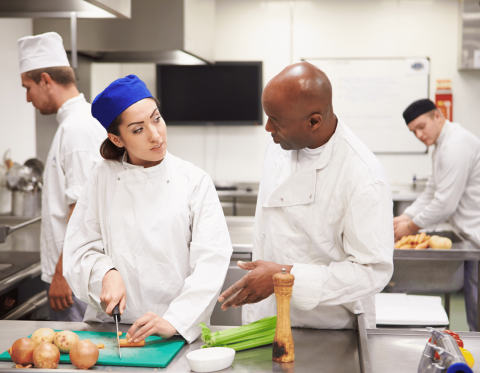 Adults in chef whites in prepping and cooking food as instructor watches