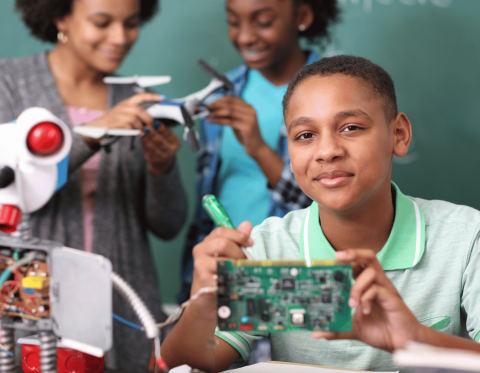 Youth holding computer circuit board with teens in the back working with robot parts